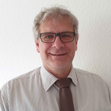 Mann mit Brille, grauen Haaren und Hemd mit Krawatte, lächelnd vor weißem Hintergrund.